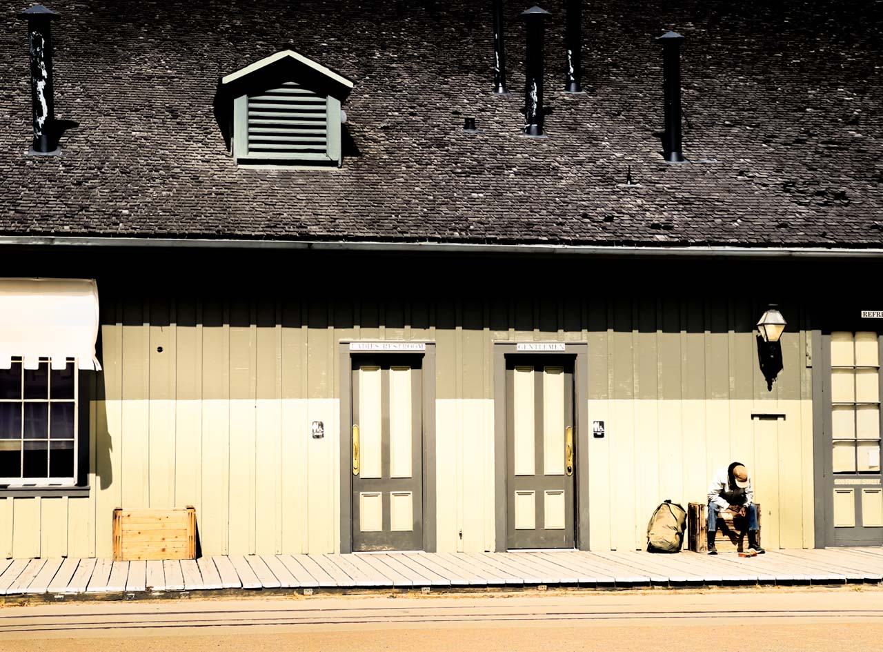 An old railroad building with a weary traveler sitting outside on the porch.